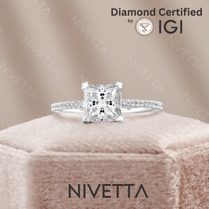 May include: A silver engagement ring with a square-shaped diamond. The diamond is set in a prong setting and is surrounded by a halo of smaller diamonds. The ring is on a pink velvet box with the word "Nivetta" printed on the front. The ring is certified by IGI.