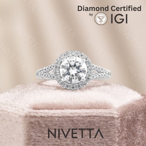 Puede incluir: Un anillo de compromiso de diamantes con engaste de halo. El anillo está hecho de oro blanco y presenta un diamante redondo brillante en el centro. El diamante está rodeado por un halo de diamantes más pequeños. El anillo está sentado en una caja de terciopelo rosa. El anillo está certificado por IGI.
