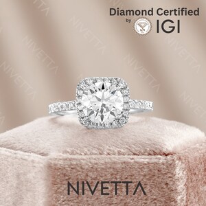 May include: A silver ring with a square-shaped diamond center stone surrounded by a halo of smaller diamonds. The ring is on a pink velvet box with the word "Nivetta" printed on the box. The diamond is certified by IGI.
