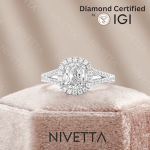May include: A silver engagement ring with a cushion-cut diamond center stone surrounded by a halo of smaller diamonds. The ring has a split shank with pave diamonds on each side. The ring is in a velvet box with the word "NIVETTA" printed on the front. The ring is certified by IGI.