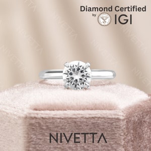 May include: A silver engagement ring with a round, clear diamond. The ring is on a pink velvet box with the word "NIVETTA" printed on the front. The diamond is certified by IGI.