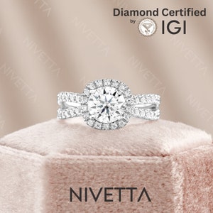 May include: A silver engagement ring with a large, round, clear diamond surrounded by a halo of smaller diamonds. The ring has a double band with smaller diamonds set into the band. The ring is sitting on a light pink velvet box. The text "Diamond Certified by IGI" is visible in the upper right corner of the image. The text "NIVETTA" is visible in the lower center of the image.