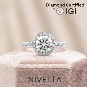 May include: A silver engagement ring with a round diamond center stone surrounded by a halo of smaller diamonds. The ring is set on a white velvet box with the word "NIVETTA" printed on the front. The diamond is certified by IGI.