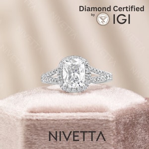 May include: A silver engagement ring with a cushion-cut diamond center stone surrounded by a halo of smaller diamonds. The ring has a split shank with pave diamonds on each side. The ring is in a pink velvet box with the word "NIVETTA" printed on the front. The ring is certified by IGI.