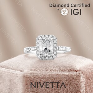 May include: A silver engagement ring with a large, square-shaped diamond set in a halo setting. The diamond is surrounded by smaller diamonds. The ring is on a pink velvet cushion. The text "Diamond Certified by IGI" is visible in the top right corner of the image.