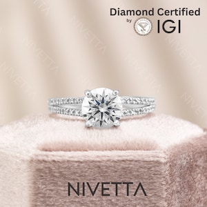 May include: A silver engagement ring with a round, clear diamond center stone and smaller diamonds set in the band. The ring is in a pink velvet box with the word "NIVETTA" printed on the front. The ring is certified by IGI.