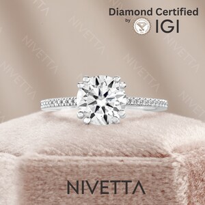 May include: A silver engagement ring with a round, clear diamond center stone and a band of smaller diamonds. The ring is in a velvet box and the words "Diamond Certified by IGI" are visible in the background.