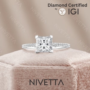 May include: A silver engagement ring with a square-shaped diamond. The diamond is set in a prong setting and is surrounded by a halo of smaller diamonds. The ring is on a pink velvet box with the word "Nivetta" printed on the front. The ring is certified by IGI.