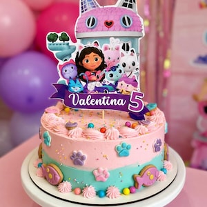 May include: A two-tiered birthday cake with pink and turquoise frosting, decorated with paw prints, sprinkles, and biscuits. A cake topper features a cartoon cat house and characters, with the name "Valentina" and the number "5".