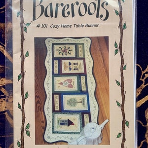 May include: A Bareroots #101 Cozy Home Table Runner sewing pattern in a clear plastic sleeve. The cover features a tan background with a brown vine border and a photograph of a rectangular table runner with five distinct quilted blocks.