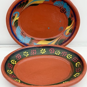 May include: Two terracotta-colored, oval-shaped serving platters. The top platter has a blue, yellow, and green floral design around the rim. The bottom platter has a green rim with yellow and pink dot patterns. Both are hand-painted.