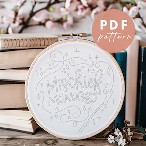 May include: A white embroidery hoop with a black and white design that says "Mischief Managed" with a wand and stars. The hoop is sitting on a stack of books. The text "PDF pattern" is in a pink circle.