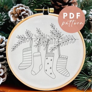 Op de afbeelding: Een zwart-wit borduurpatroon van vier kousen die aan een tak met bladeren hangen. Het patroon is op een witte achtergrond en is ingelijst in een houten hoepel. De tekst "PDF pattern" staat op een roze cirkel in de rechterbovenhoek van de afbeelding.