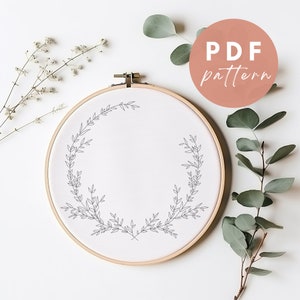 May include: A white embroidery hoop with a simple line drawing of a wreath of leaves. The hoop is on a white background with sprigs of white flowers and eucalyptus leaves.