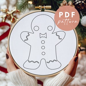May include: A white embroidery hoop with a black line drawing of a gingerbread person wearing a bow tie. The text "PDF pattern" is on a pink circle in the upper right corner of the image.