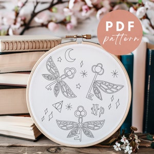 Puede incluir: Un aro de bordado blanco con un dibujo de línea negra de tres llaves con alas, una luna creciente, estrellas y los símbolos de las Reliquias de la Muerte y Harry Potter. El aro está sobre una pila de libros. El texto "PDF pattern" está en un círculo rosa en la esquina superior derecha de la imagen.