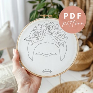 May include: A white embroidery hoop with a black line drawing of a woman's face with a flower crown. The text "PDF pattern" is on a pink circle in the upper right corner of the image.