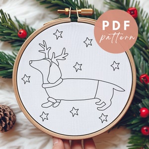 May include: A white embroidery hoop with a black line drawing of a dachshund wearing a reindeer costume and surrounded by stars. The hoop is labeled "PDF pattern".