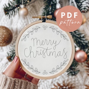 Könnte beinhalten: Ein weißer Stickrahmen mit dem Text "Merry Christmas" in schwarzer Kursivschrift. Der Rahmen ist von einem einfachen Kranz aus Blättern und Beeren umgeben.