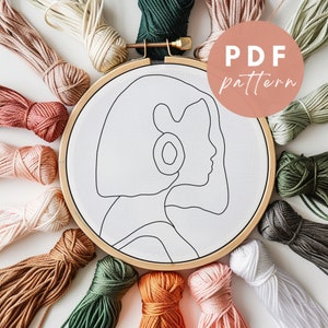 May include: A wooden embroidery hoop with white fabric inside, featuring a line drawing of a woman's face. The hoop is surrounded by a variety of colorful embroidery floss.