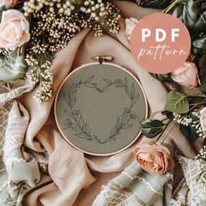 Peut inclure: Un cerceau de broderie vert avec un motif de cœur floral noir. Le cerceau est entouré de fleurs roses et blanches et de verdure. Le texte "PDF pattern" est écrit dans un cercle sur le côté droit de l'image.