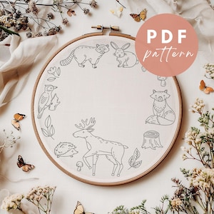 Könnte beinhalten: Ein weißer Stickrahmen mit einer Schwarzweiß-Zeichnung von Waldtieren und Pflanzen. Die Tiere umfassen einen Waschbär, ein Kaninchen, einen Fuchs, ein Reh, einen Specht und einen Igel. Die Pflanzen umfassen Blätter, einen Pilz und einen Baumstumpf. Der Text "PDF pattern" ist in einem rosa Kreis oben rechts im Bild geschrieben.
