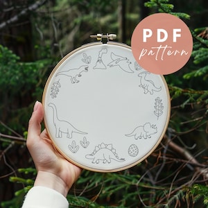May include: A white embroidery hoop with a black line drawing of dinosaurs and a volcano. The hoop is held in a hand against a green background. The text "PDF pattern" is on a pink circle in the upper right corner of the image.