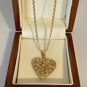 May include: A gold heart-shaped locket necklace with a delicate filigree design. The necklace is displayed in a wooden box.