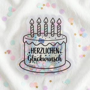 Puede incluir: Un recipiente transparente en forma de pastel con cinco ilustraciones de velas en la parte superior. El recipiente está lleno de dinero y confeti. El texto "HERZLICHEN Glückwunsch" está escrito en la parte delantera. Fondo blanco con confeti.