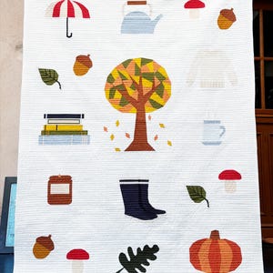 May include: White quilt featuring autumn-themed illustrations. The design includes a tree, umbrella, acorns, leaves, a teapot, a sweater, a mug, a jar, boots, a mushroom, a stack of books, and a pumpkin. The quilt has a textured surface.