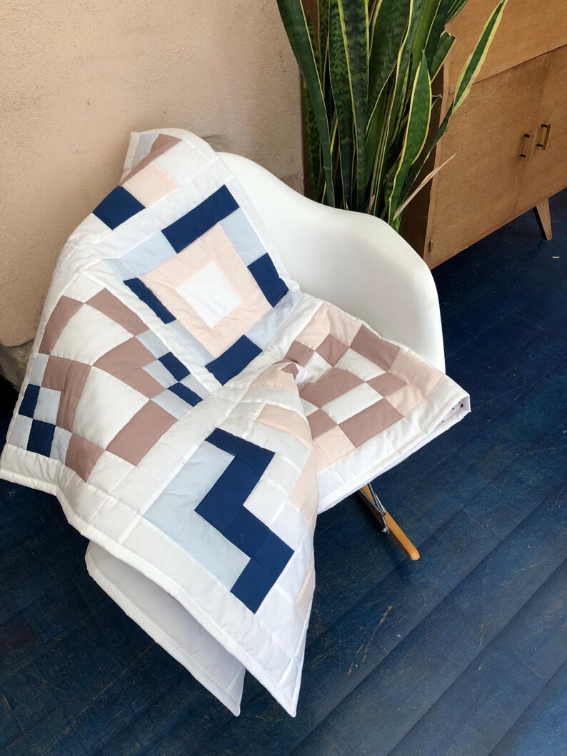 May include: A white quilt with a geometric pattern in navy, blush, and brown, draped over a white chair. The quilt features a square design with alternating blocks of colour and a zig-zag pattern.