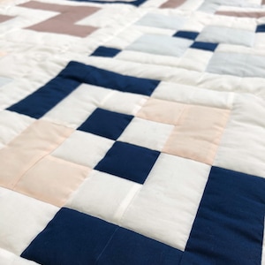 May include: A close-up of a handmade quilt with a geometric pattern. The quilt features squares in white, navy blue, light pink, and light blue. The quilt is made of fabric and has a quilted texture.