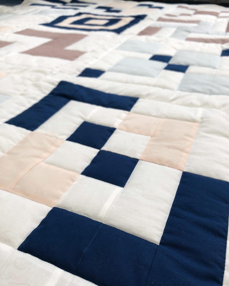 May include: A close-up of a handmade quilt with a geometric pattern. The quilt features squares in white, navy blue, light pink, and light blue. The quilt is made of fabric and has a quilted texture.