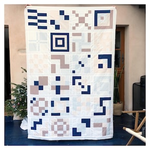 May include: A handmade quilt with a geometric pattern in shades of white, blue, and taupe. The quilt features a grid design with various square and rectangular shapes. The quilt is held up, showcasing its size and the detailed stitching.