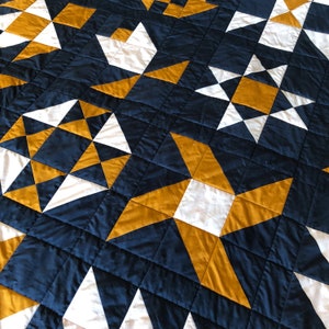 May include: A quilt made of navy blue, white, and gold fabric. The quilt is made up of a repeating pattern of stars and squares.