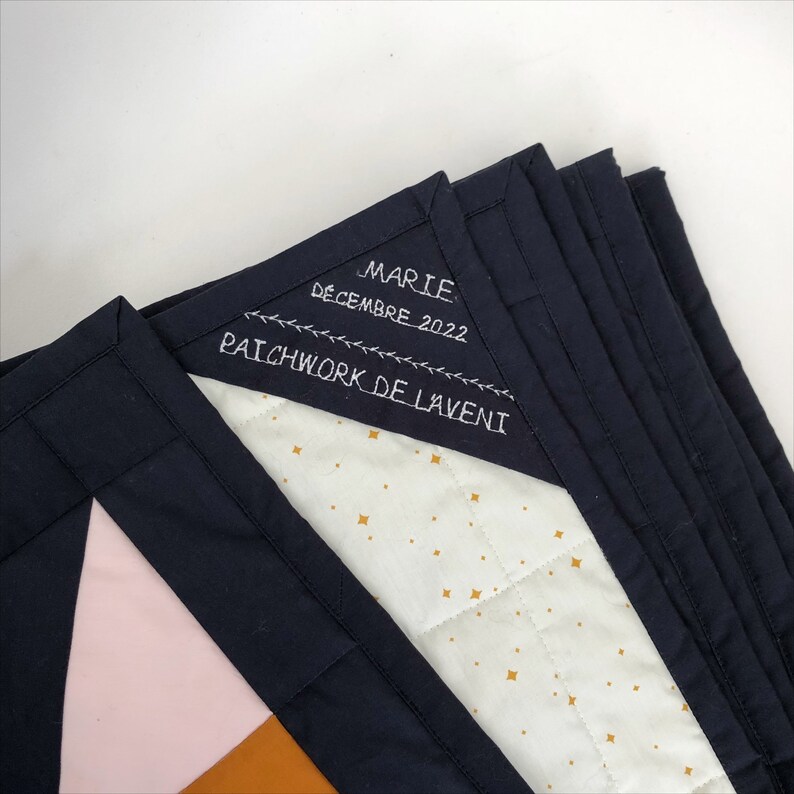 May include: A black quilted fabric with a white background and gold stars. The fabric has a label sewn on it that says "Marie Decembre 2022 Patchwork de Laveni".