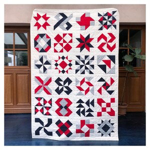 Może przedstawiać: Biała kołdra z patchworkowym wzorem z czerwonymi i szarymi geometrycznymi kształtami. Kołdra składa się z 25 pojedynczych kwadrat&oacute;w, każdy z innym wzorem.