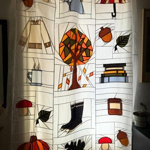 May include: White fabric curtain with a patchwork design featuring autumn-themed illustrations. The design includes a tree, acorns, leaves, a teapot, a pumpkin, boots, and a mushroom. The color palette includes orange, brown, and black.