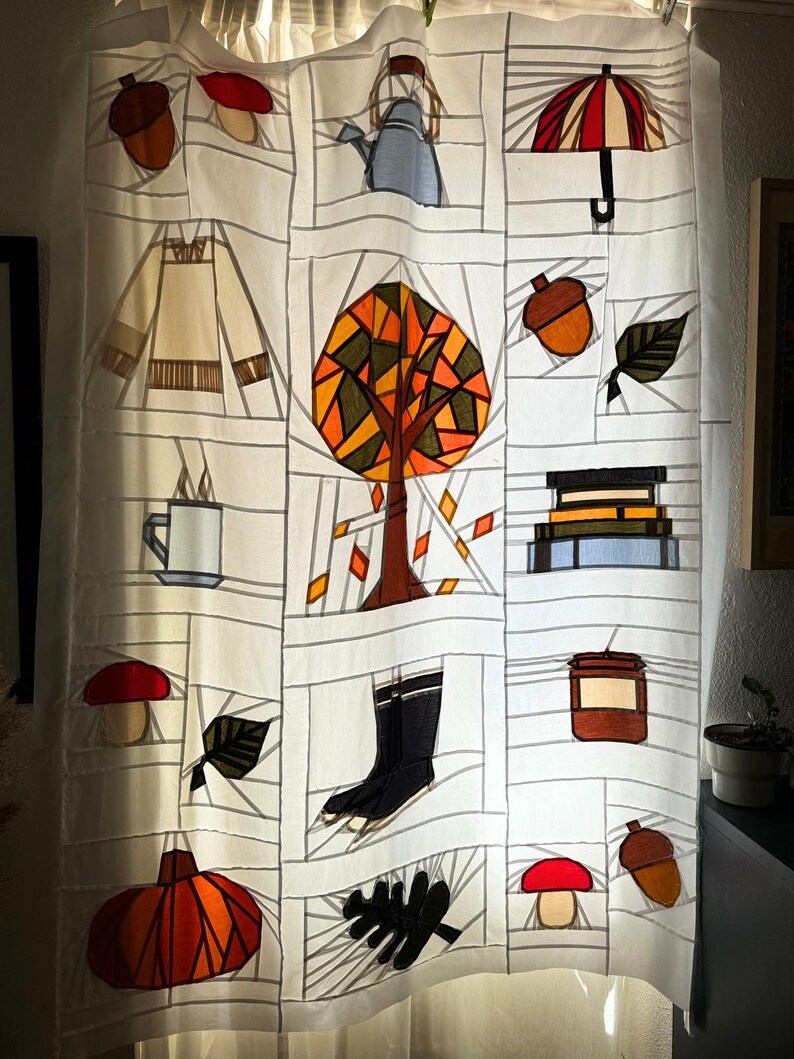 May include: White fabric curtain with a patchwork design featuring autumn-themed illustrations. The design includes a tree, acorns, leaves, a teapot, a pumpkin, boots, and a mushroom. The color palette includes orange, brown, and black.