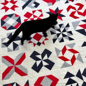 Może przedstawiać: Czarny kot chodzi po białej kołdrze z czerwonymi, szarymi i granatowymi geometrycznymi wzorami. Kołdra jest wykonana z kwadratowych kawałk&oacute;w materiału zszytych ze sobą, tworząc patchworkowy wz&oacute;r.