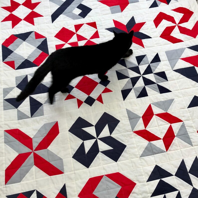 Może przedstawiać: Czarny kot chodzi po białej kołdrze z czerwonymi, szarymi i granatowymi geometrycznymi wzorami. Kołdra jest wykonana z kwadratowych kawałk&oacute;w materiału zszytych ze sobą, tworząc patchworkowy wz&oacute;r.