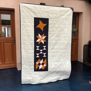 May include: A white quilt with a geometric pattern in shades of orange, pink, and navy blue. The pattern is a vertical row of four different shapes, including a star, a starburst, a series of triangles, and a diamond.