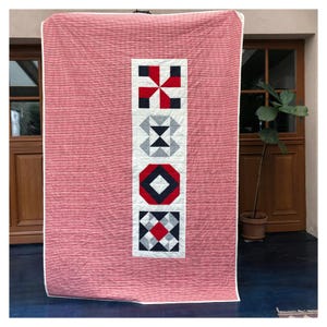 Może przedstawiać: Kołdra w kratę w kolorze czerwonym i białym z pionowym panelem pięciu r&oacute;żnych geometrycznych blok&oacute;w patchworkowych w kolorze czarnym, białym, czerwonym i szarym.