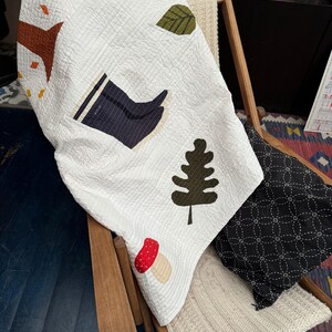 May include: White quilted blanket with appliqued autumn-themed designs, including a tree, leaves, a boot, a mushroom, and a tree. The blanket is draped over a wooden chair with a black patterned pillow and a cream-colored knitted blanket.
