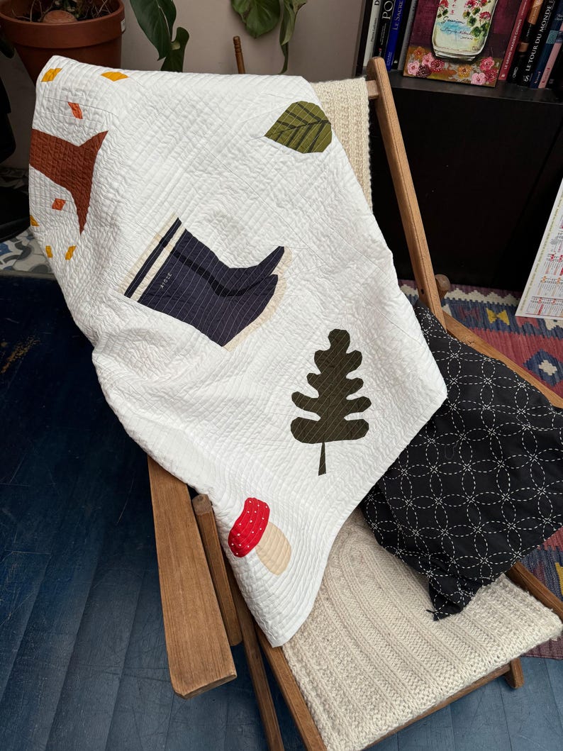 May include: White quilted blanket with appliqued autumn-themed designs, including a tree, leaves, a boot, a mushroom, and a tree. The blanket is draped over a wooden chair with a black patterned pillow and a cream-colored knitted blanket.
