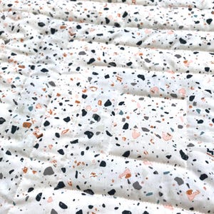 May include: Close-up of a white quilted fabric with a terrazzo pattern. The pattern features small, irregular shapes in black, grey, peach, and brown. The quilting creates parallel lines across the fabric.