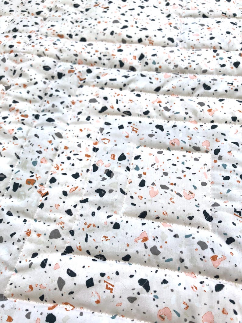 May include: Close-up of a white quilted fabric with a terrazzo pattern. The pattern features small, irregular shapes in black, grey, peach, and brown. The quilting creates parallel lines across the fabric.