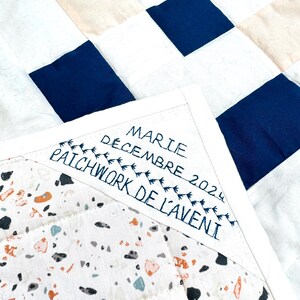 May include: A close-up of a handmade quilt with a patchwork design. The quilt features squares of white, navy blue, and beige fabric. The corner displays embroidered text: "MARIE DECEMBRE 2024 PATCHWORK DE L'AVENIR."