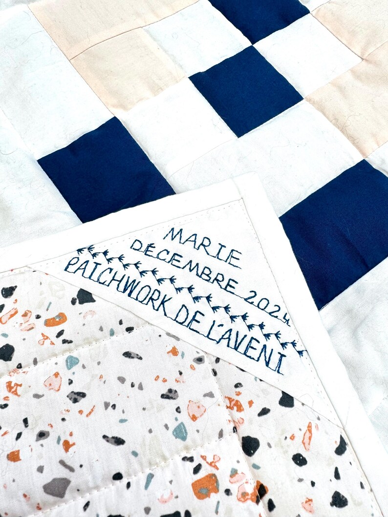 May include: A close-up of a handmade quilt with a patchwork design. The quilt features squares of white, navy blue, and beige fabric. The corner displays embroidered text: "MARIE DECEMBRE 2024 PATCHWORK DE L'AVENIR."