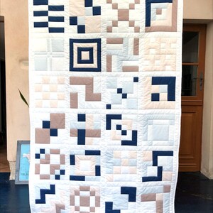 May include: A white quilt with a geometric pattern in navy blue, pale blue, and beige. The quilt has a grid of squares and rectangles, creating a modern, minimalist design. The quilt is displayed against a neutral background.